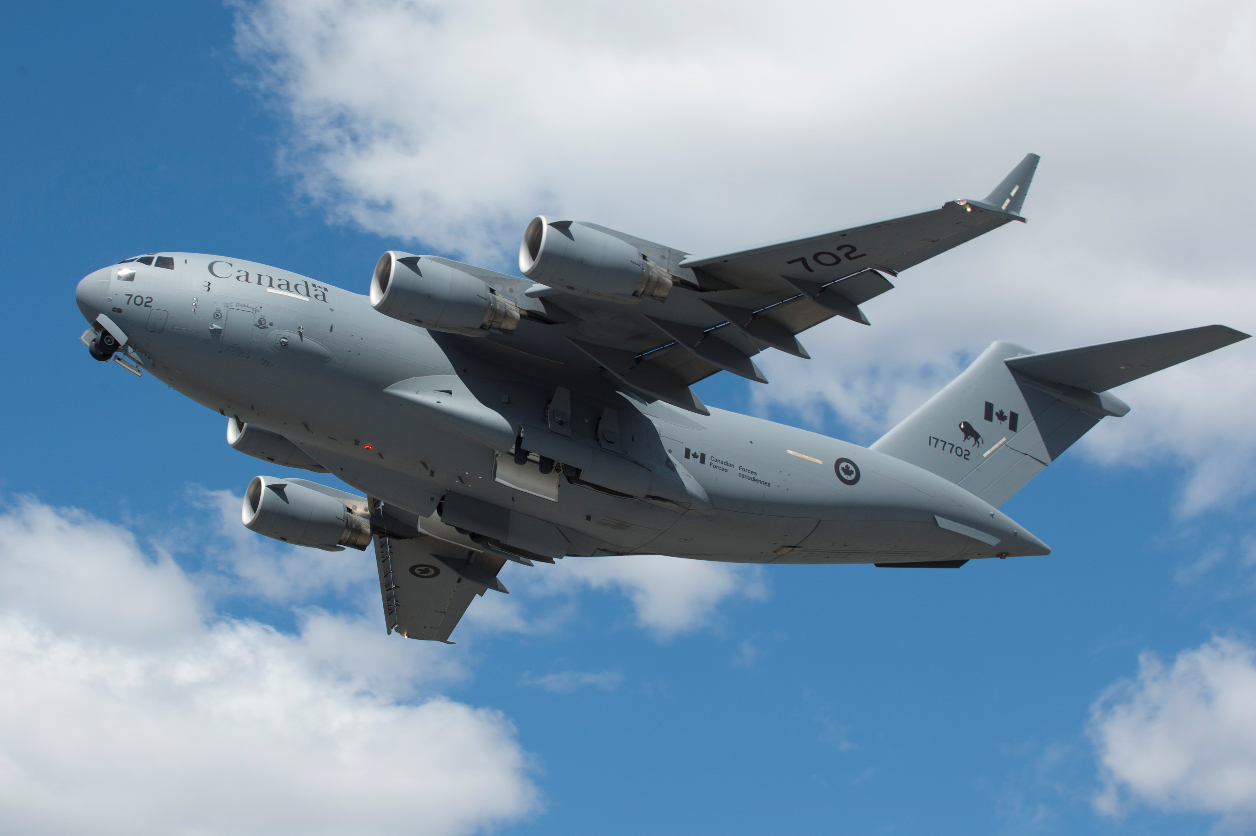 A large grey aircraft with military markings flies against a blue sky. 